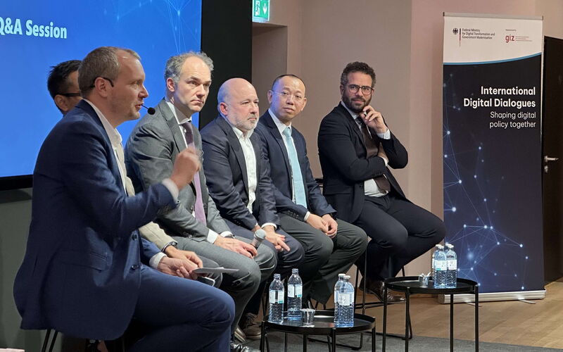 Fünf Diskussionsteilnehmende sitzen auf der Bühne und blicken nach rechts zum Moderator, der am rechten Ende sitzt und die Einführung zur Podiumsdiskussion hält.