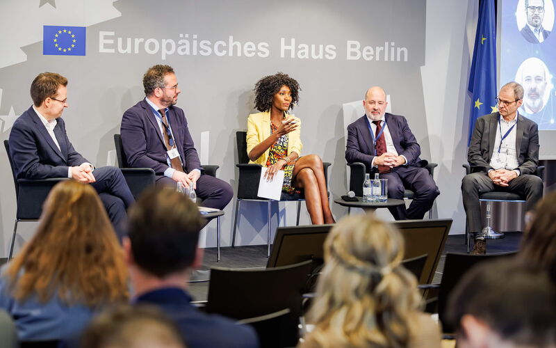 Vier Diskussionsteilnehmer und der Moderator sitzen in einem Halbkreis auf der Bühne, und der Moderator in der Mitte hält die Einführungsrede zur Veranstaltung.