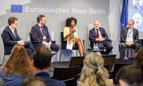 Four Panelists, plus the moderator sit on stage in a semicircle and the moderator at the center is doing the intro to the event.
