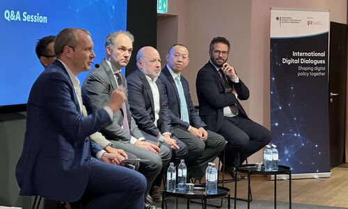 Fünf Diskussionsteilnehmende sitzen auf der Bühne und blicken nach rechts zum Moderator, der am rechten Ende sitzt und die Einführung zur Podiumsdiskussion hält.