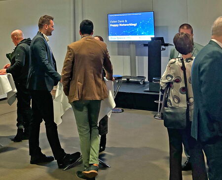 Attendees stand around high tables during a networking session at an event. A screen in the background displays the message “Vielen Dank & Happy Networking!” next to a podium. People are engaged in conversation in a modern indoor setting.