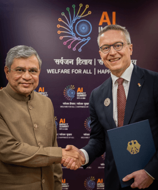 Two men in formal attire shake hands at an official meeting in front of a backdrop reading ‘AI Impact Summit.’