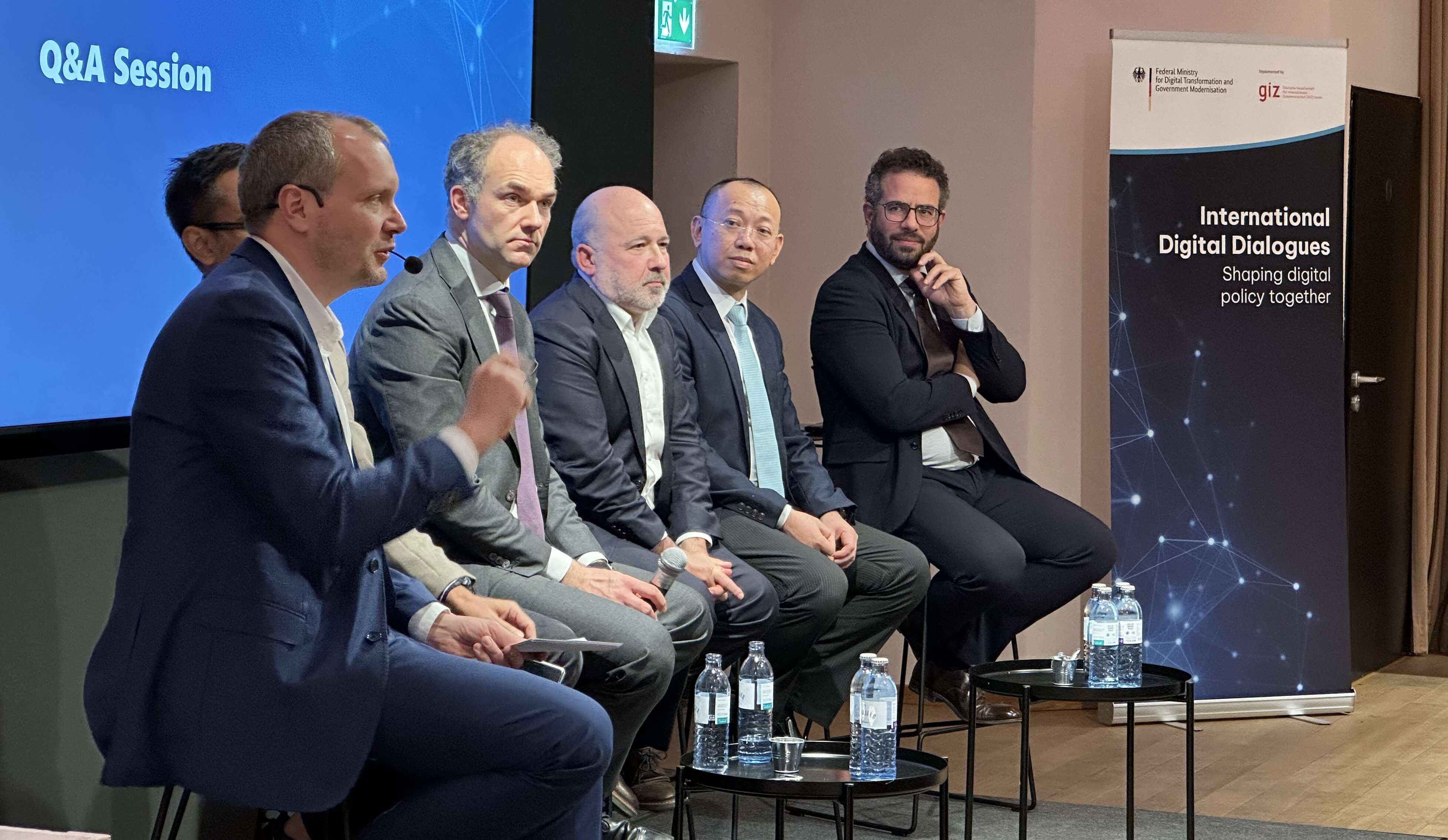 Fünf Diskussionsteilnehmer sitzen auf der Bühne und blicken nach rechts zu dem Moderator, der am rechten Ende sitzt und die Einführung zur Podiumsdiskussion hält.