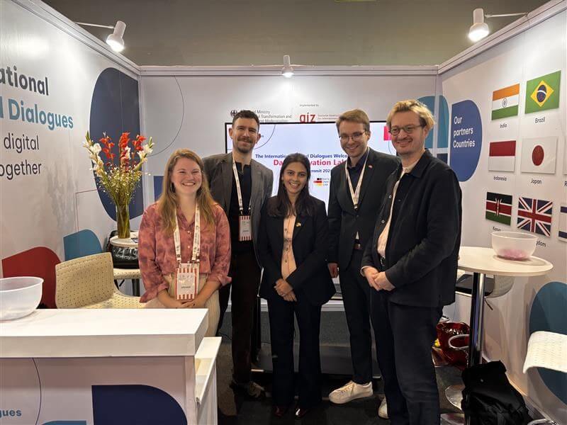 Five people stand together smiling at the International Digital Dialogues booth at the India AI Impact Summit, surrounded by informational displays and partner country flags.
